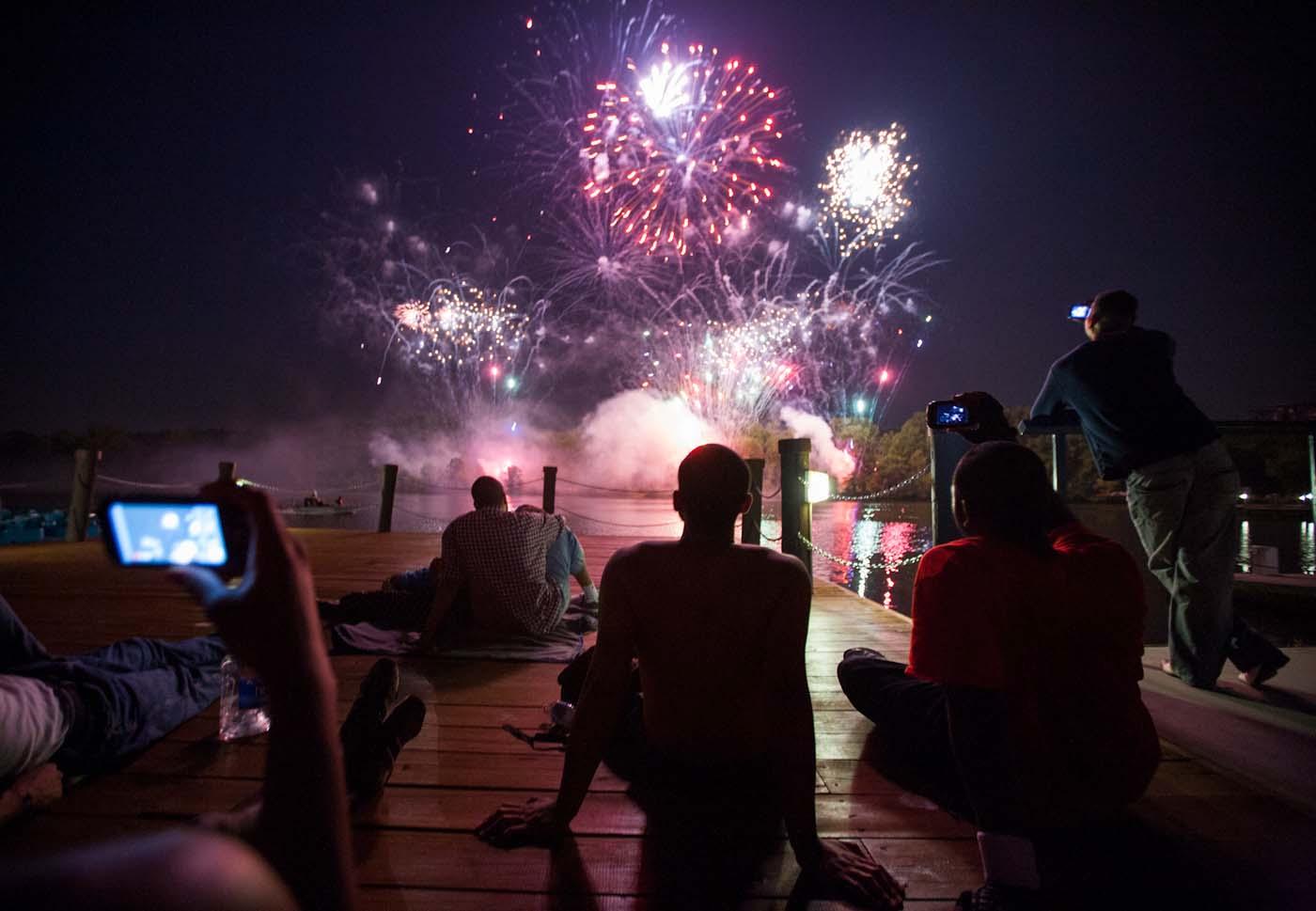 Close-up (from back) of individuals viewing fireworks from boat dock at night