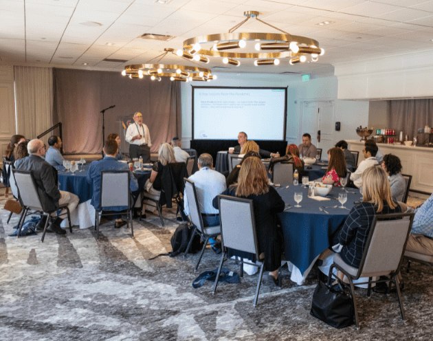 Board meeting at banquet center with two circular gold metal chandeliers, a speaker at front to left of lecturer screen, attendees look on from circular tables with gray chairs and blue tablecloths