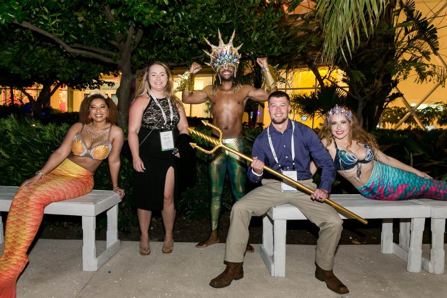 Two event attendees stand at night outside with three individuals dressed as mermaids, merman; one event attendee holds trident
