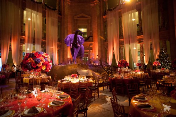 Opulent event venue inside a large hall with tall columns, featuring a giant purple elephant statue surrounded by dramatic red draping and multiple dining tables set for a gala